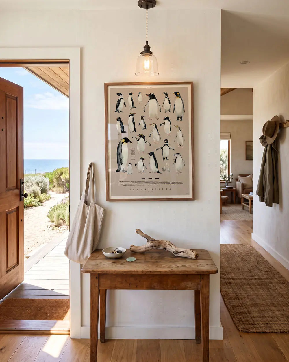 An entryway to a seaside home with a small wooden table. There is a framed artwork above the table featuring the world's penguin species.