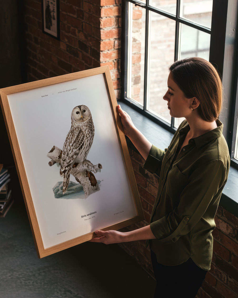 Woman holding a framed print of an owl in a room with brick walls and large windows.