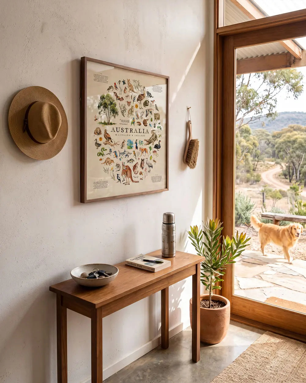 A framed art print hangs on a textured white wall in a rustic entryway. The artwork features Australian animals arranged in a circle shape. A wooden console table sits below, styled with a hat, thermos, and small bowl, next to a potted plant.