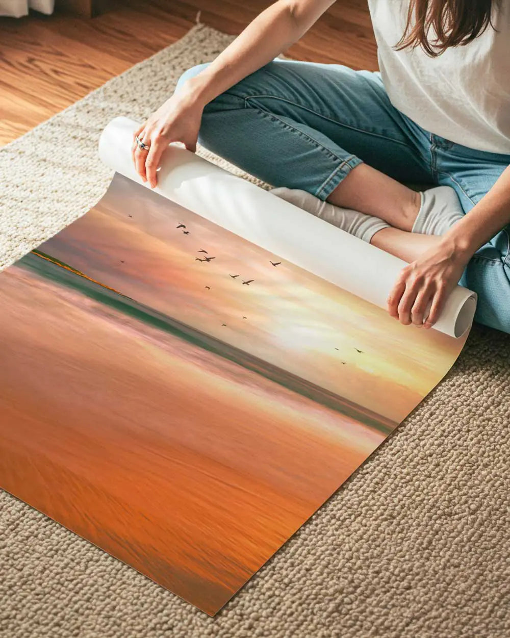A woman sits cross-legged on a carpet unrolling a large beach scene poster.