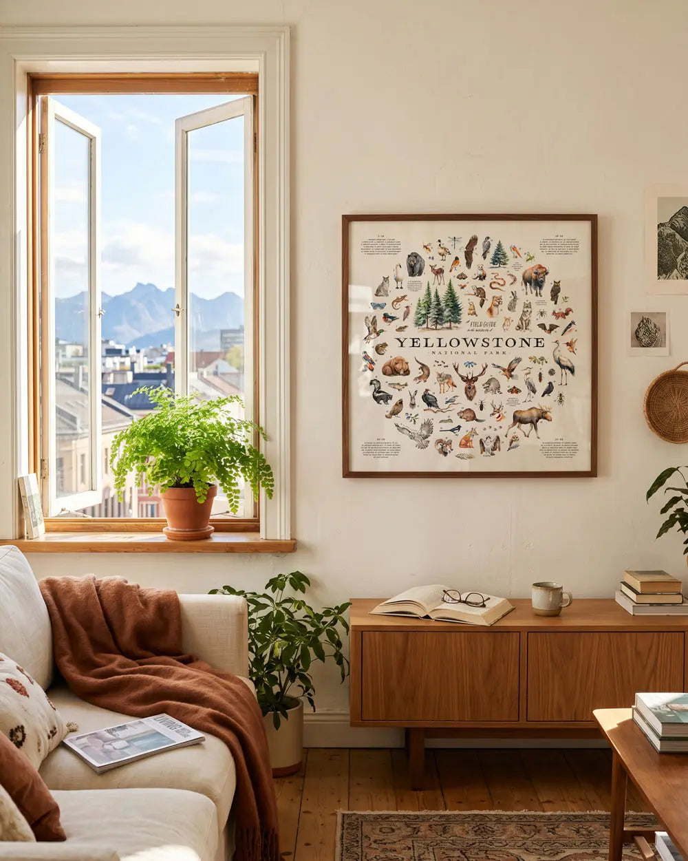 A framed Yellowstone National Park nature art print hangs on a cream wall in a cozy, sunlit living room. The print features a detailed vintage-style illustration of various animals and plants of the park.
